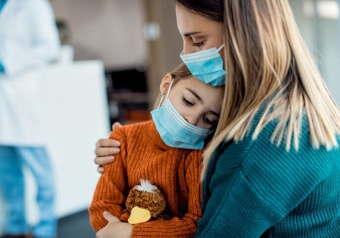 Mother comforting her sad daughter while sitting in waiting room in the hospital and wearing protective face masks.